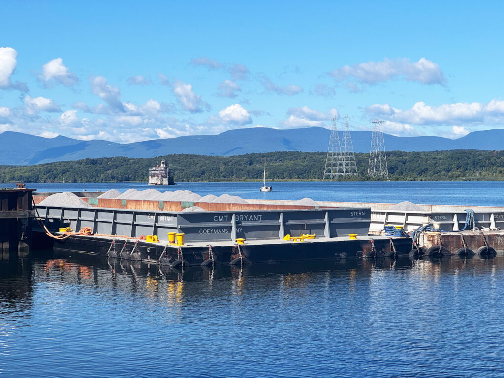 Stone barges at the Colarusso dock complex, waiting to be shipped on the river. In the background the Hudson-Athens Lighthouse, August 2025. Photo by Annik LaFarge, author of Composing Olana